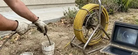 Plumber feeding a sewer camera cable into a cleanout beside a monitor and reel