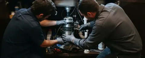 Two plumbers installing a garbage disposal under a kitchen sink